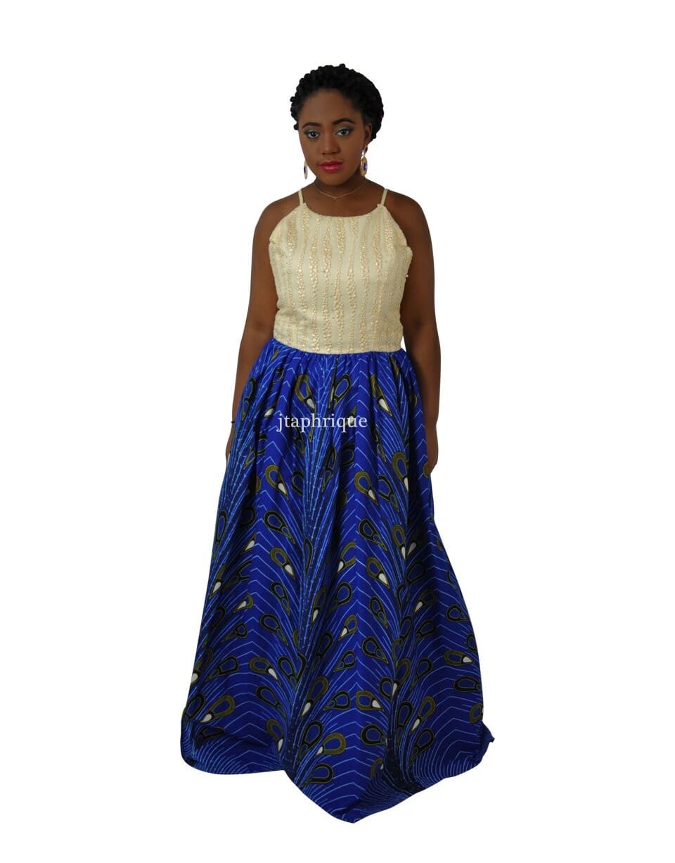 Frontal of model wearing a special occasion dress with a cream spaghetti strap top embellished with sequins and beads. Has a full dark blue maxi skirt with all over African Ankara feather or peacock print.