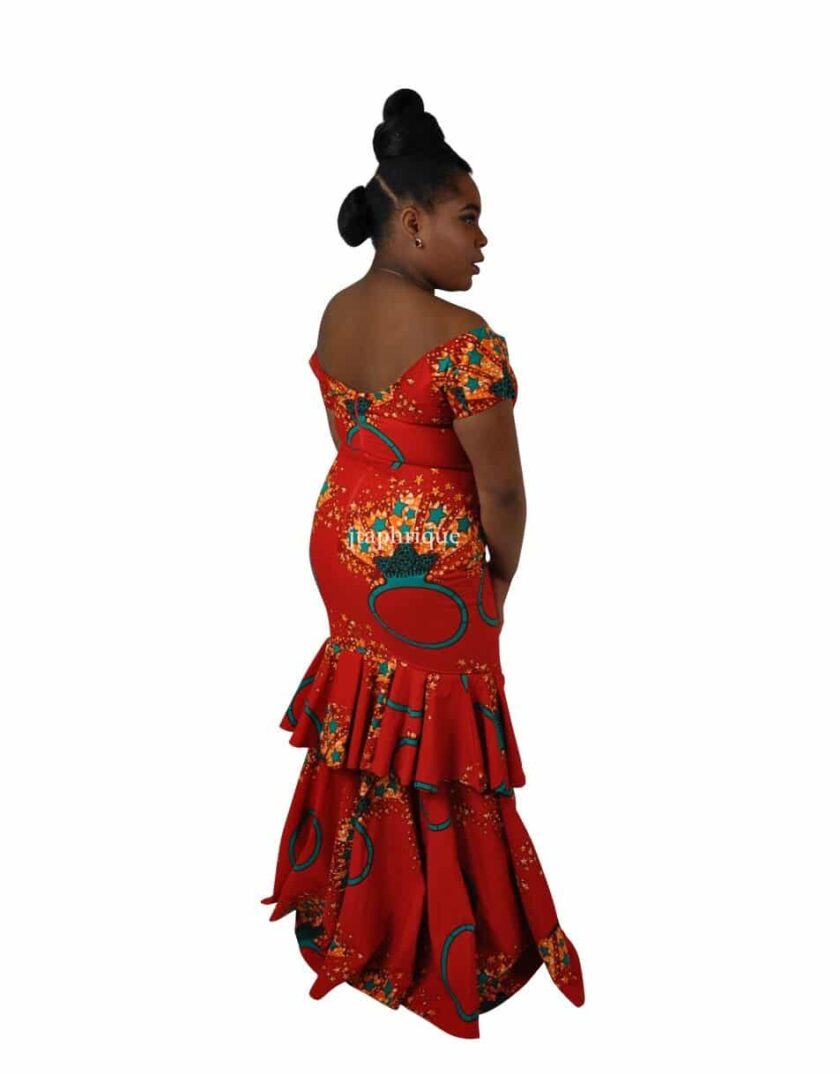 Back shot of model wearing a red off shoulder long fit and flare occasion dress with sweetheart neckline, front slit, peplum ruffles on the back in all over Ankara ring and star print.