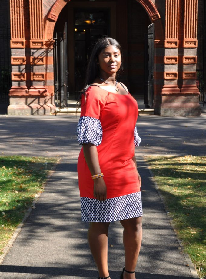 Frontal of model wearing a pencil dress in solid red polished Cotton and checker pattern Ankara print detail on sleeves and hem.