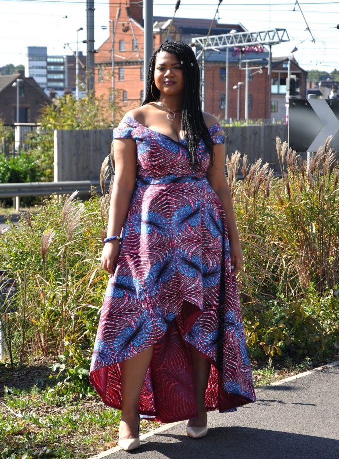 Frontal of model wearing a red and blue African inspired high low dress in all over feather Ankara print.
