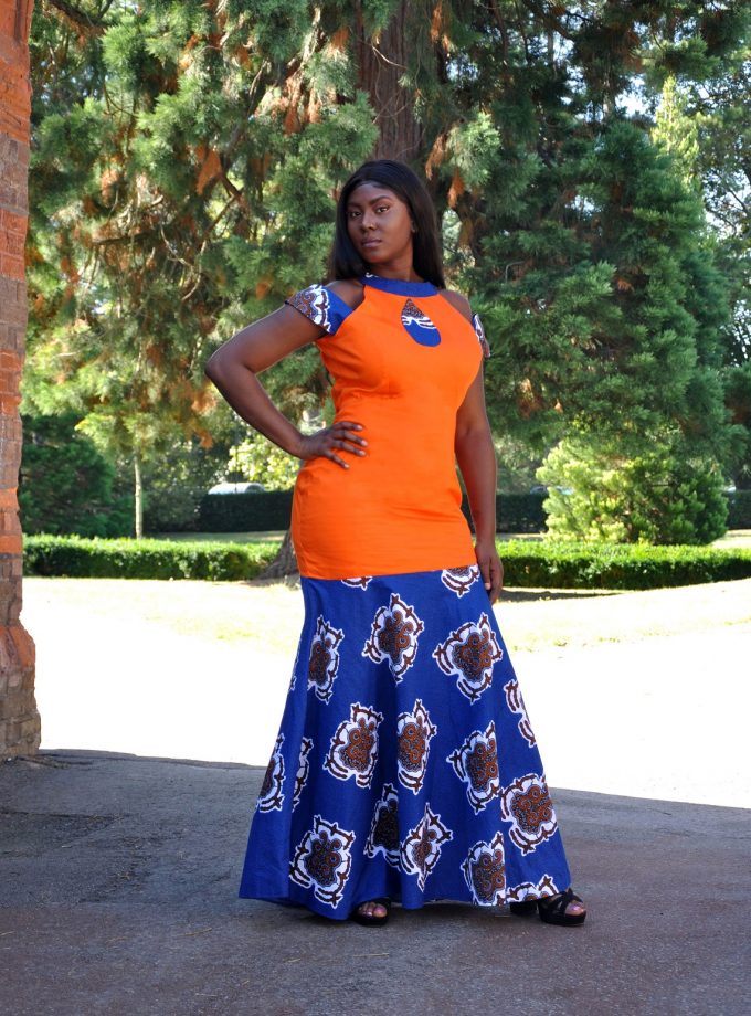 Frontal of model wearing an orange fit and flare occasion dress with blue African Ankara print pattern detail on hem from thigh.