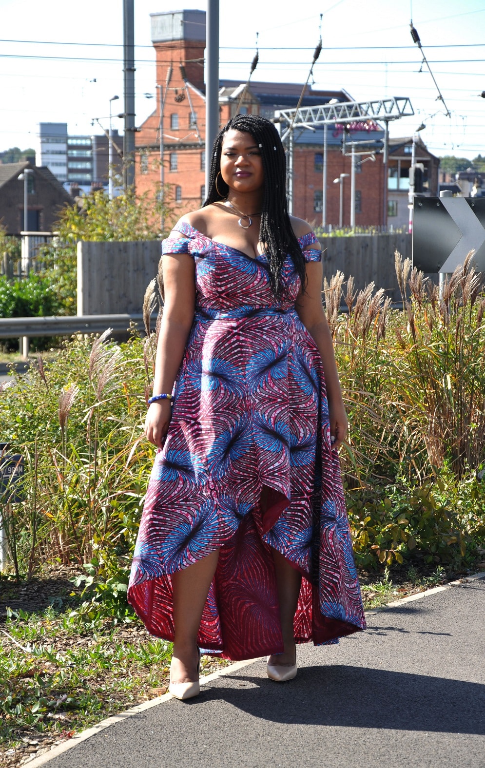 Frontal of model wearing a red and blue African inspired high low off shoulder special occasion dress in all over feather Ankara print.