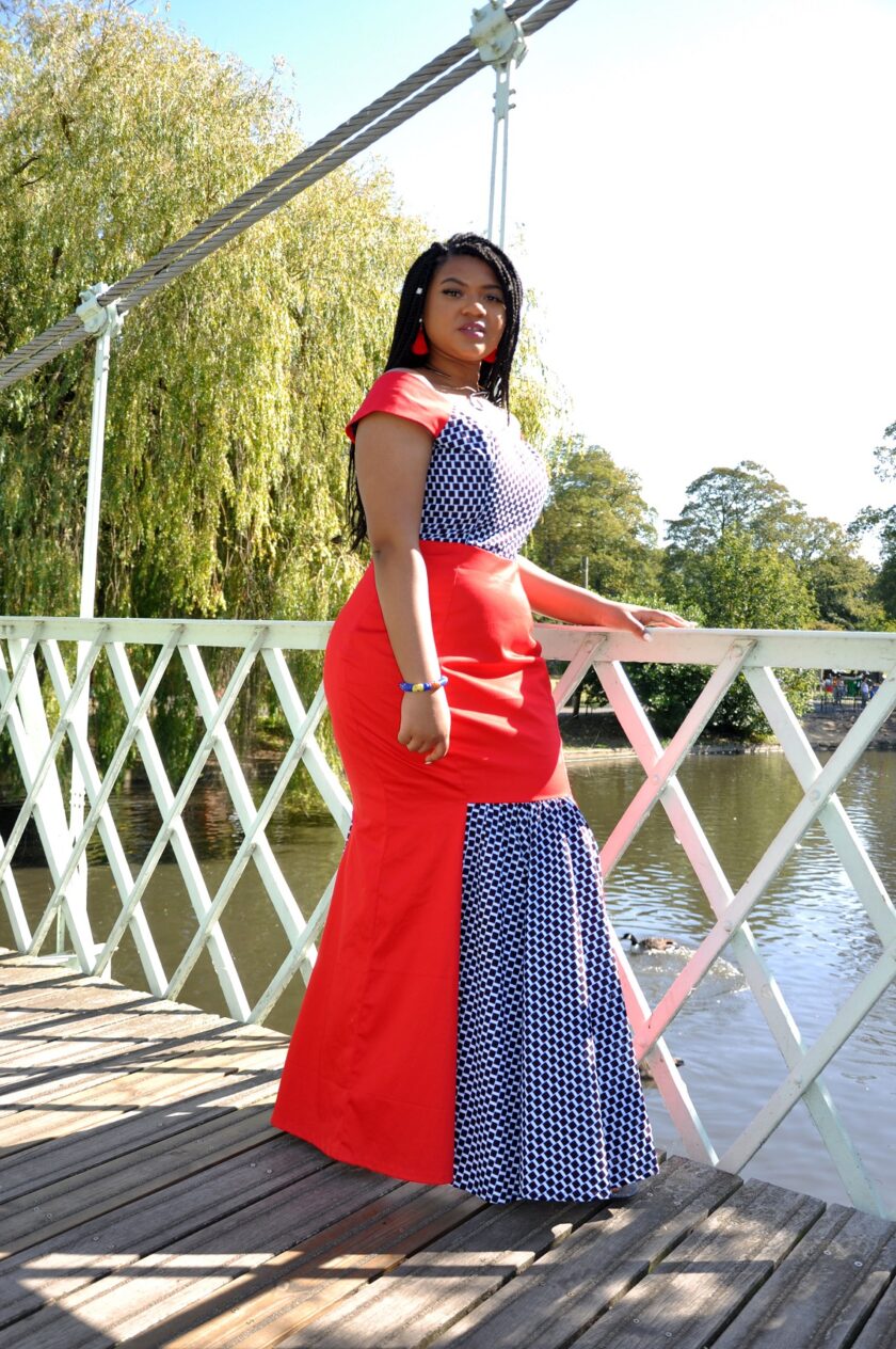 Side shot of model wearing a red special occasion fit and flare dress with contrasting African Ankara checker pattern on top and pleated fishtail.
