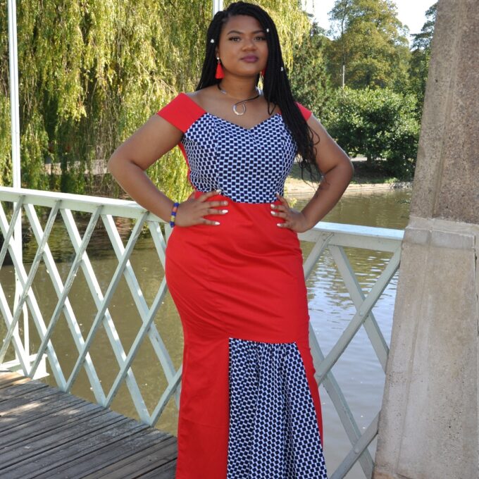 Frontal of model wearing a red special occasion fit and flare dress with contrasting African Ankara checker pattern on top and pleats.
