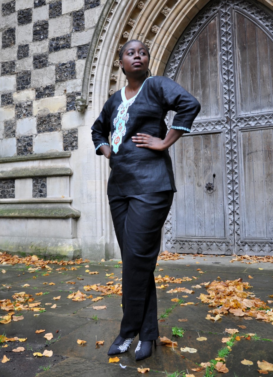 Frontal of model wearing a ladies black traditional African linen trouser suit / pantsuit with cream, turquoise and orange embroidery on the front, neckline, back and sleeve trim.
