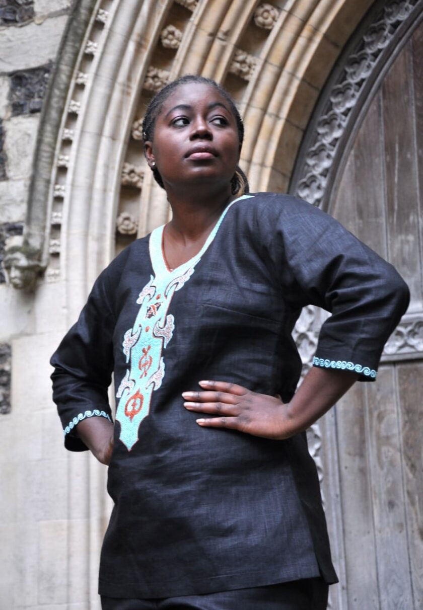 Side shot of model wearing a ladies black traditional African linen trouser suit / pantsuit with cream, turquoise and orange embroidery on the front, neckline, back and sleeve trim.