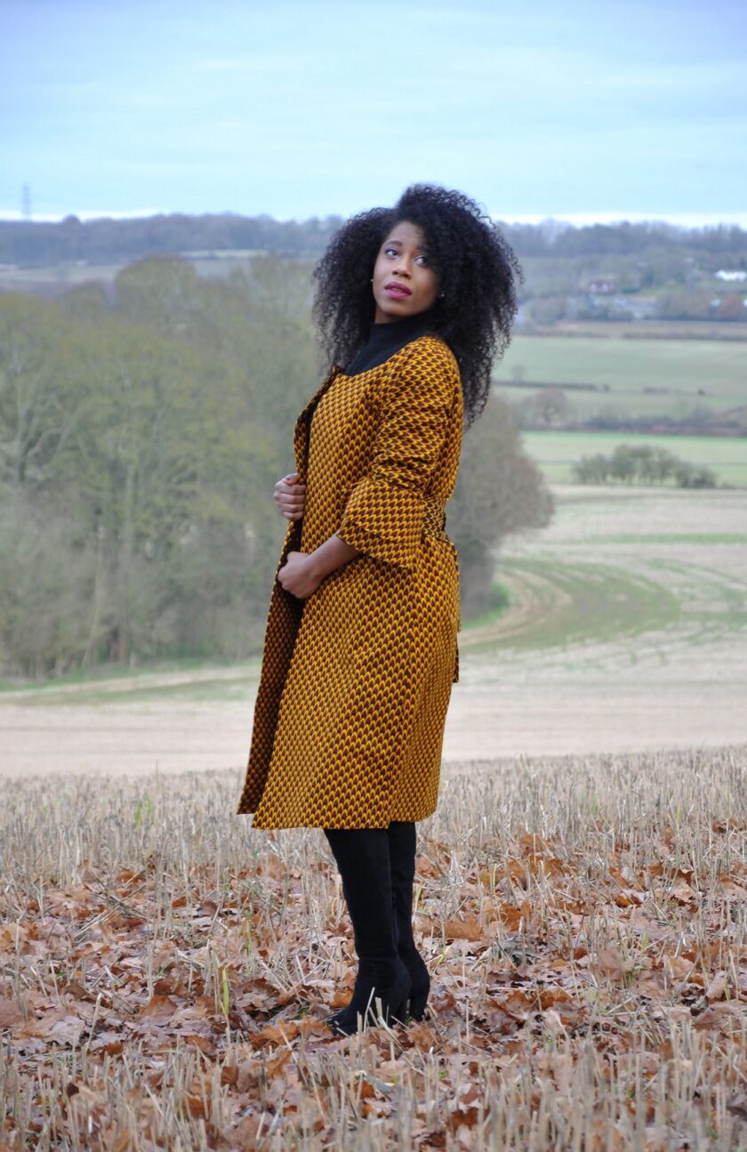 Side shot of model wearing a ladies mustard yellow ruffle sleeve belted trench coat in all over brown African print.