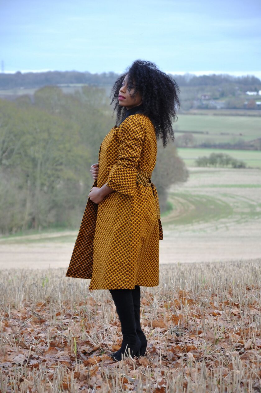 Side shot of model wearing a ladies mustard yellow ruffle sleeve belted trench coat in all over brown African print.