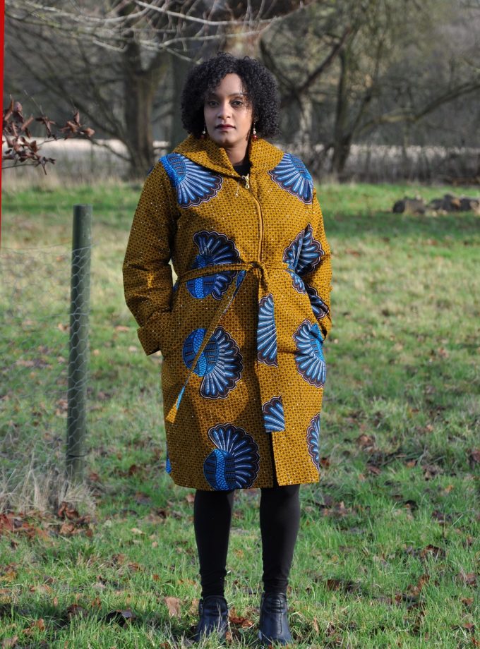 Full frontal of model wearing a mustard yellow ladies trench coat in all over blue African shell print.