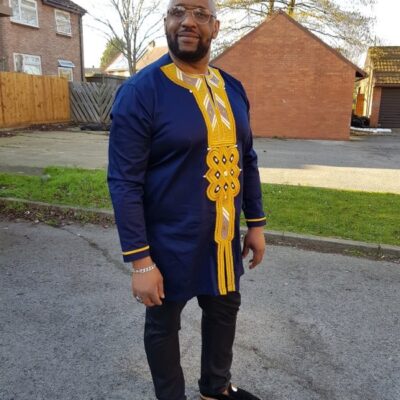 Full frontal of model wearing a men's navy blue longline shirt embroidered with gold African embroidery detail on neckline, chest and cuffs.