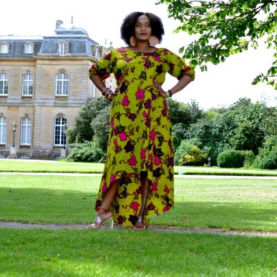 Frontal of model wearing a lime green long dress with half sleeves, high-low asymmetrical hem in all over pink, purple and green floral African print pattern.
