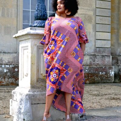 Frontal of model wearing a long pink dress with flared half sleeves, an asymmetrical high-low hem in all over red, blue and gold African print pattern.