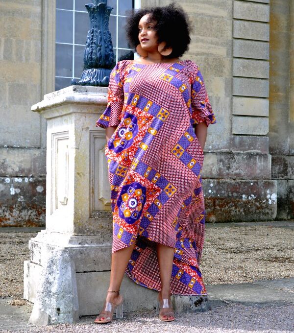 Frontal of model wearing a long pink dress with flared half sleeves, an asymmetrical high-low hem in all over red, blue and gold African print pattern.