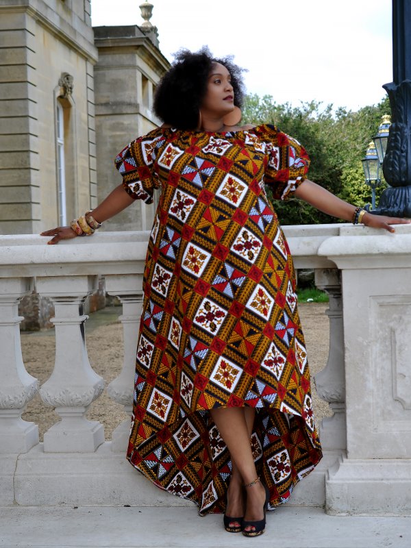 Frontal of model wearing a gold coloured maxi dress with high-low asymmetrical hem, off shoulder neckline, short puff sleeves in all over multi-coloured African print pattern.