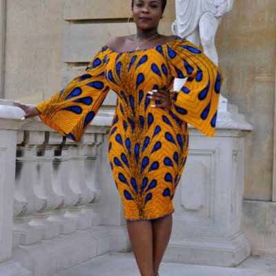 Frontal of model wearing a glamorous yellow / gold African-inspired knee-length off shoulder bodycon dress with bell sleeve in all over blue Ankara feather or peacock print pattern.