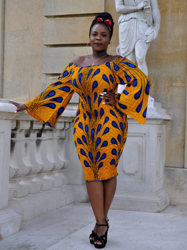 Frontal of model wearing a glamorous yellow / gold African-inspired knee-length off shoulder bodycon dress with bell sleeve in all over blue Ankara feather or peacock print pattern.