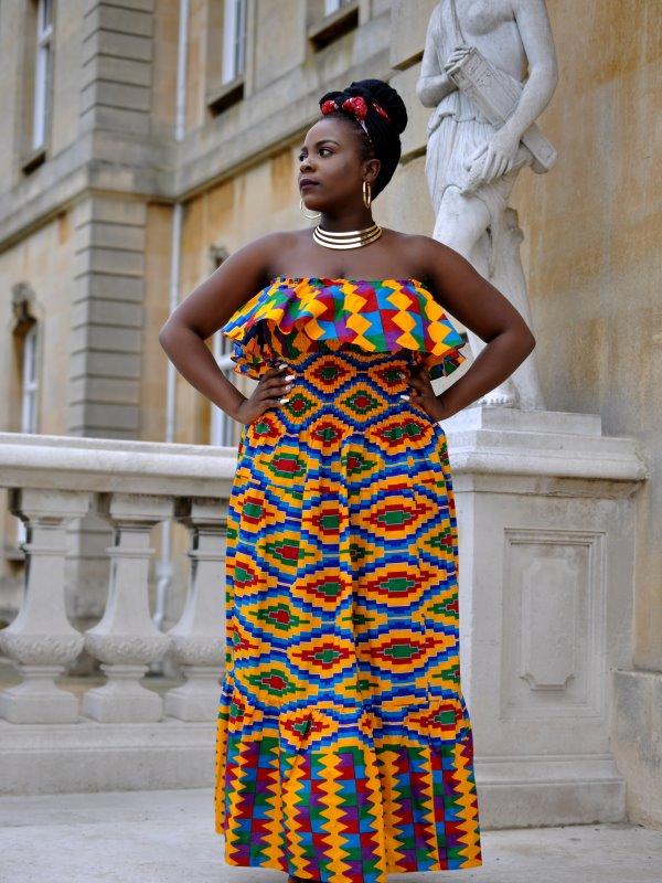 Full frontal of model wearing a colourful strapless maxi dress with peplum frill on the neckline, a shirred waistline in yellow, blue, green all over African Kente print pattern.
