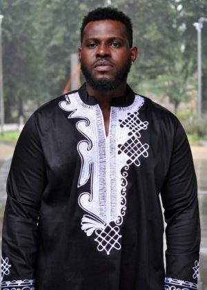 Frontal of model wearing a men's black traditional African shirt with intricate white embroidery on the chest.