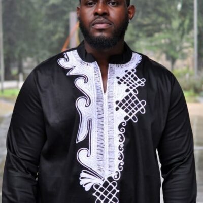 Frontal of model wearing a men's black traditional African shirt with intricate white embroidery on the chest.