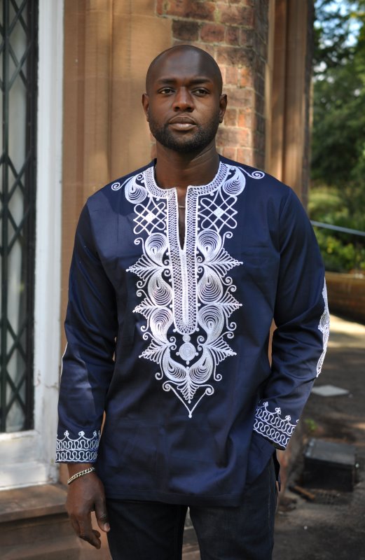 Frontal of model wearing a navy blue polished cotton shirt with silver African embroidery on the front in an intricate royal style pattern.