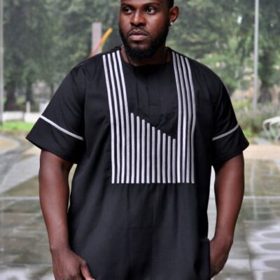 Frontal of model wearing a black short sleeved traditional African shirt with silver striped embroidery on the front and sleeves.
