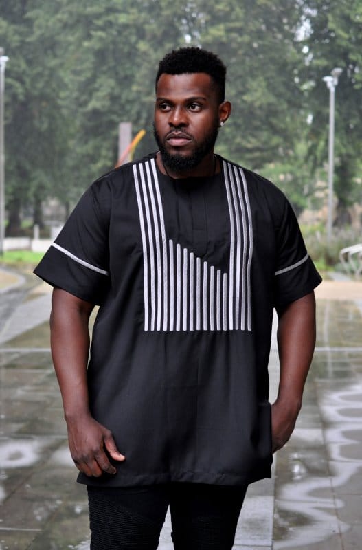 Frontal of model wearing a black short sleeved traditional African shirt with silver striped embroidery on the front and sleeves.