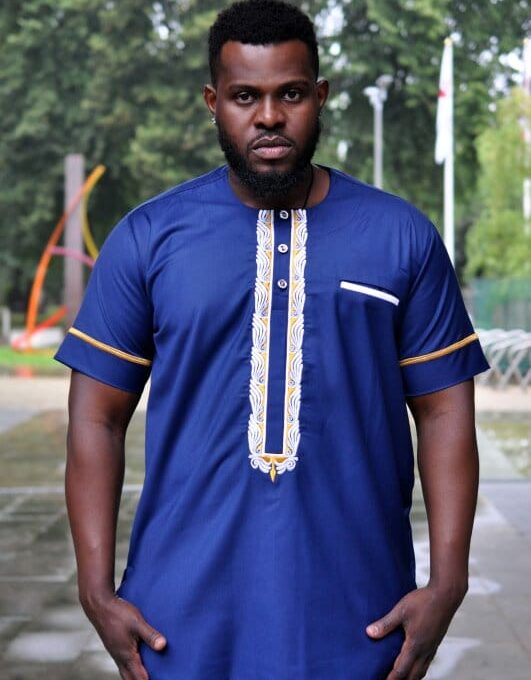 Frontal of model wearing a short sleeve blue African shirt with white and gold panel embroidery detail on the chest and gold embroidery detail on the sleeves.