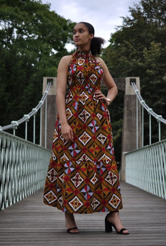 Frontal of model wearing a gold or mustard yellow halterneck summer maxi dress in all over African Ankara print.