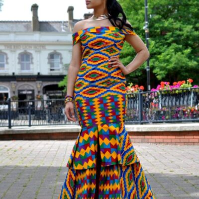 Full frontal of model wearing a short sleeve, off shoulder, fit and flare dress in all over vibrant multi-coloured African print. Features a ruffle hem, flared from the thigh with two tiered ruffles.