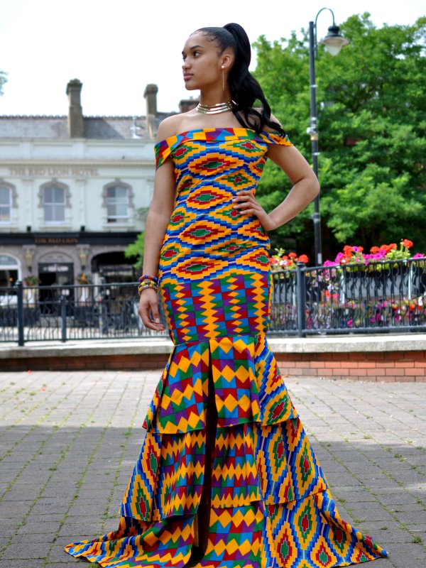 Full frontal of model wearing a short sleeve, off shoulder, fit and flare dress in all over vibrant multi-coloured African print. Features a ruffle hem, flared from the thigh with two tiered ruffles.