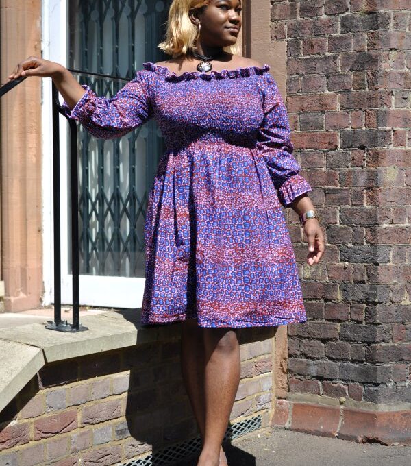 Full frontal of model wearing a purple knee-length boho dress in all over African print, with a shirred bodice.