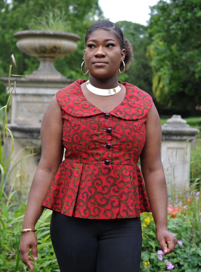 Close frontal of model wearing a sleeveless red peplum top with gold swirly African print all over.