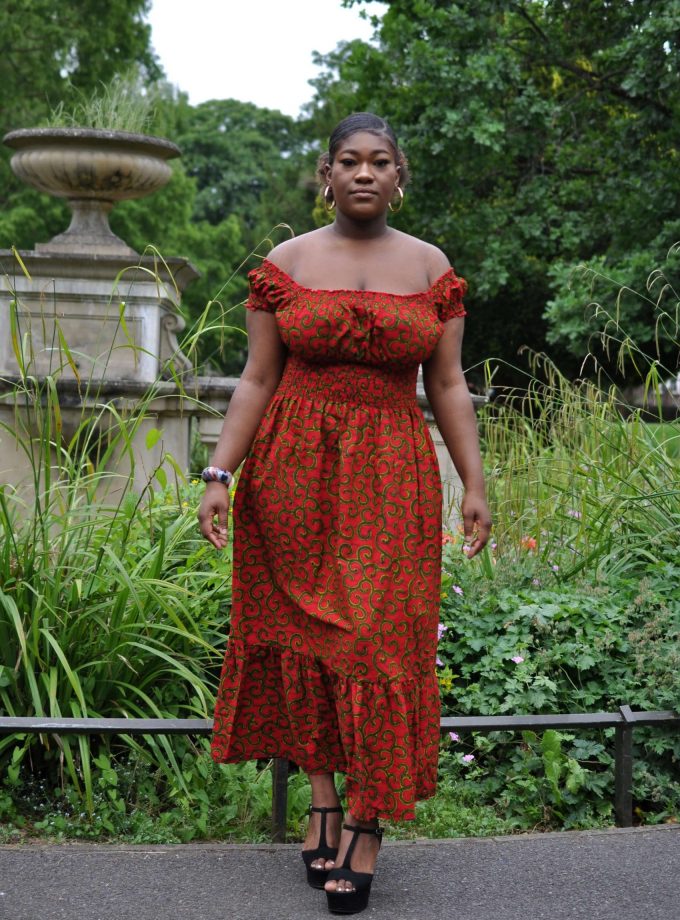 Full frontal of model wearing a red and gold African print maxi dress with shirred waist and all over swirly print.
