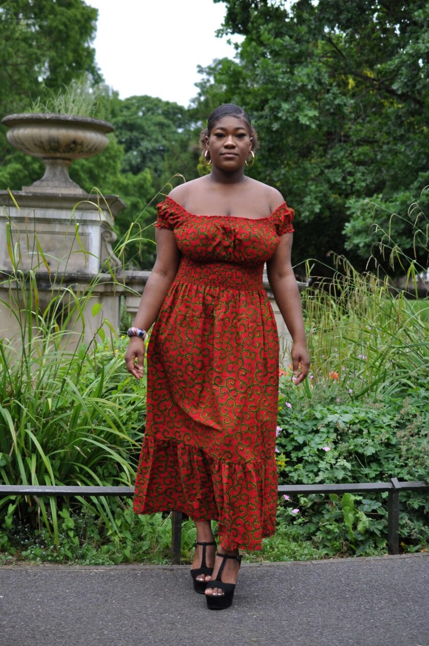 Full frontal of model wearing a red and gold African print maxi dress with shirred waist and all over swirly print.