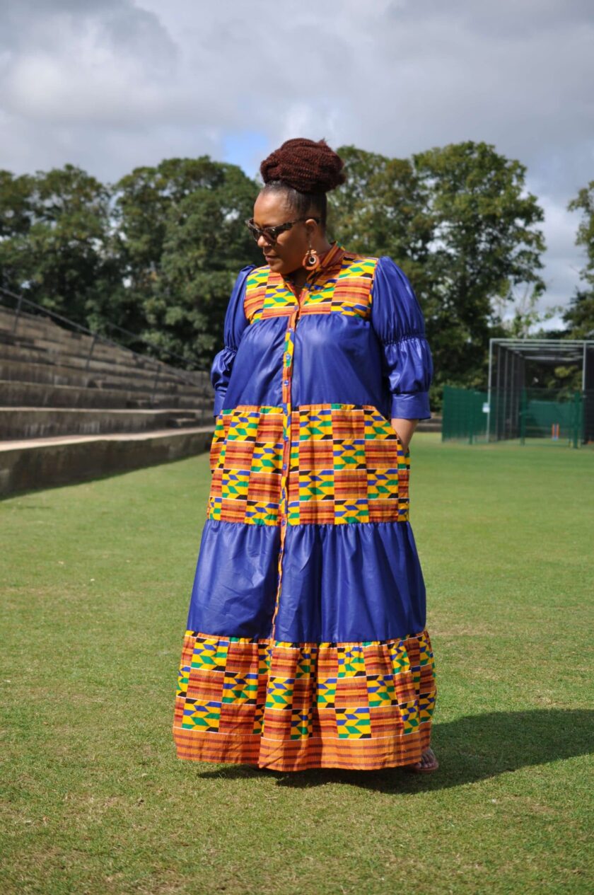 Frontal of model wearing a long blue and Kente print smock dress with stand collar and lantern sleeves.