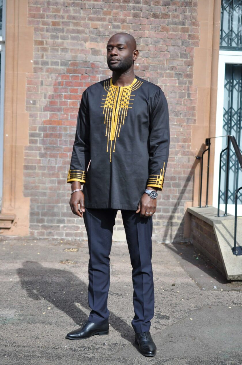 Frontal of model wearing a men's traditional African black shirt with gold embroidery on the neckline and cuffs.