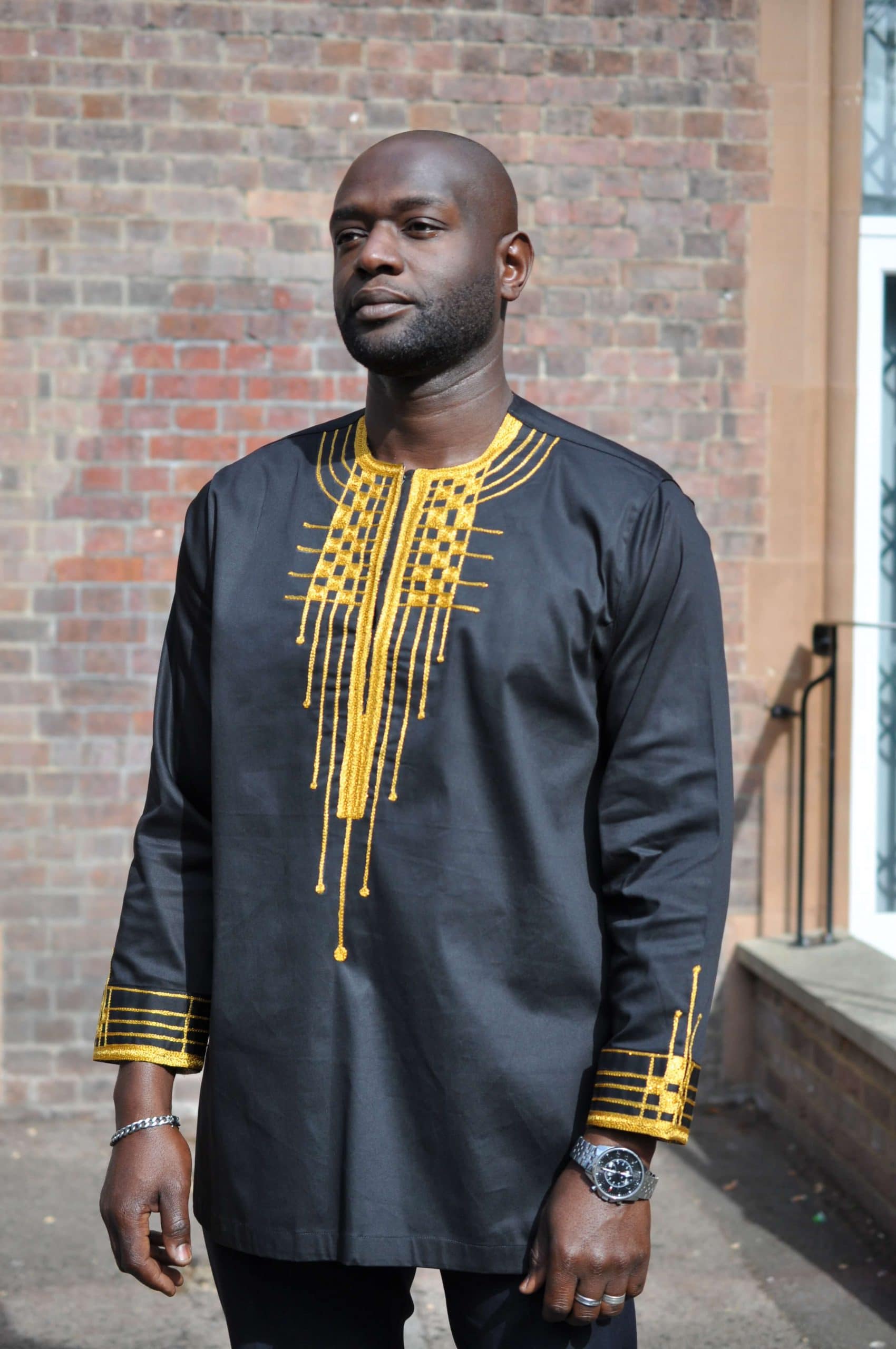 Frontal of model wearing a men's traditional African black shirt with gold embroidery on the neckline and cuffs.