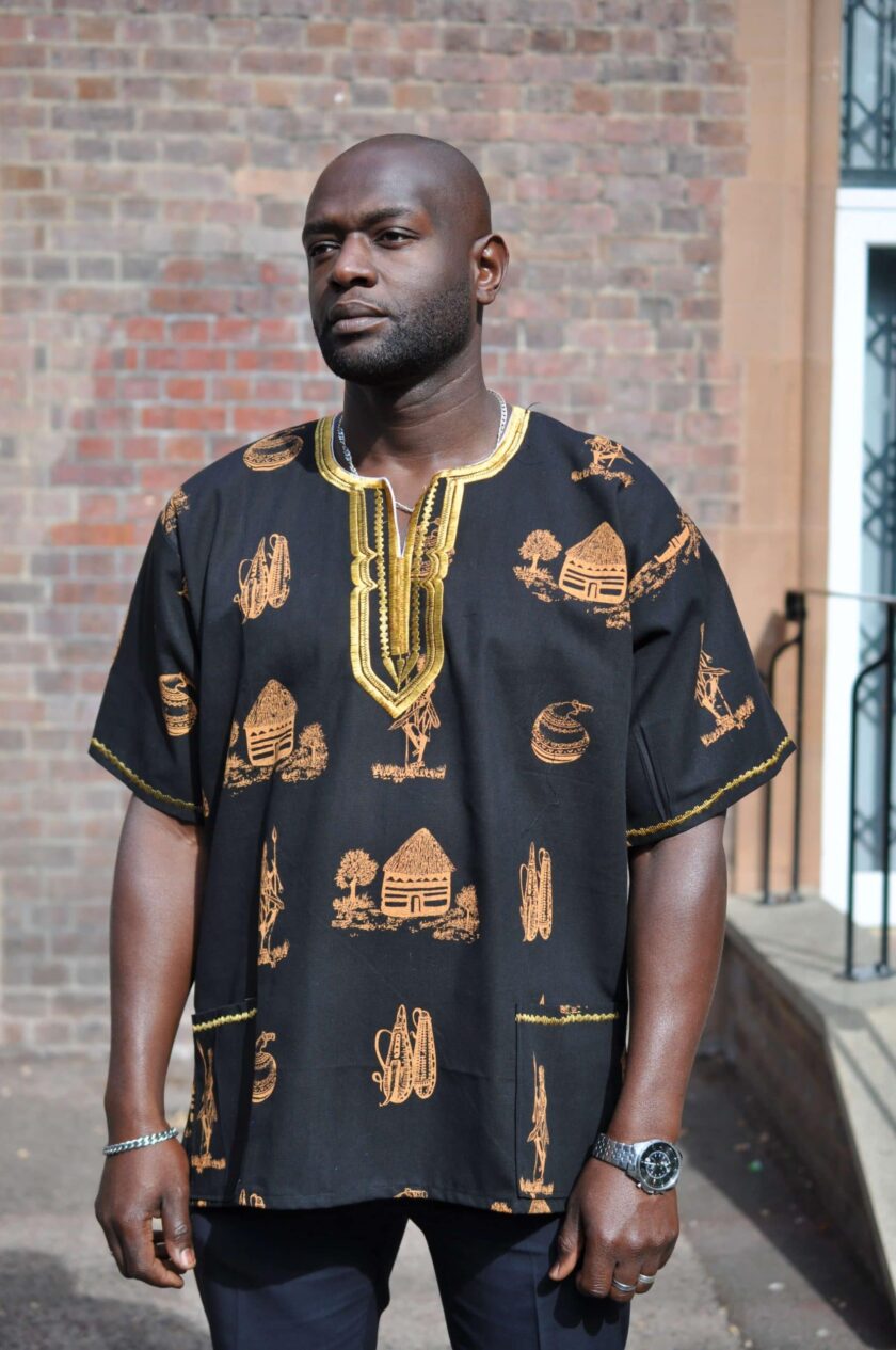 Frontal of model wearing a men's black short sleeve shirt with gold embroidery on the neckline and traditional Kenya scenes and artefacts in an all over pattern.
