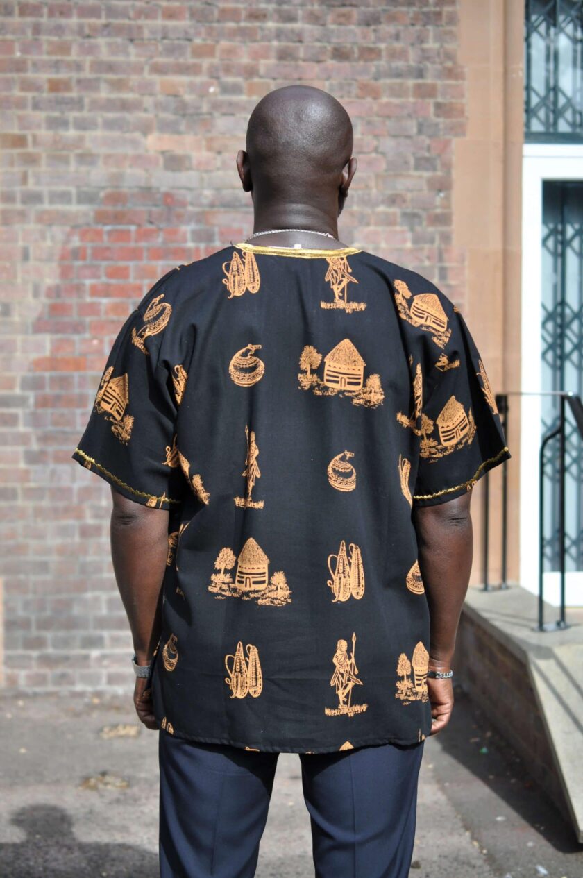 Back shot of model wearing a men's black short sleeve shirt with gold embroidery on the neckline and traditional Kenya scenes and artefacts in an all over pattern.