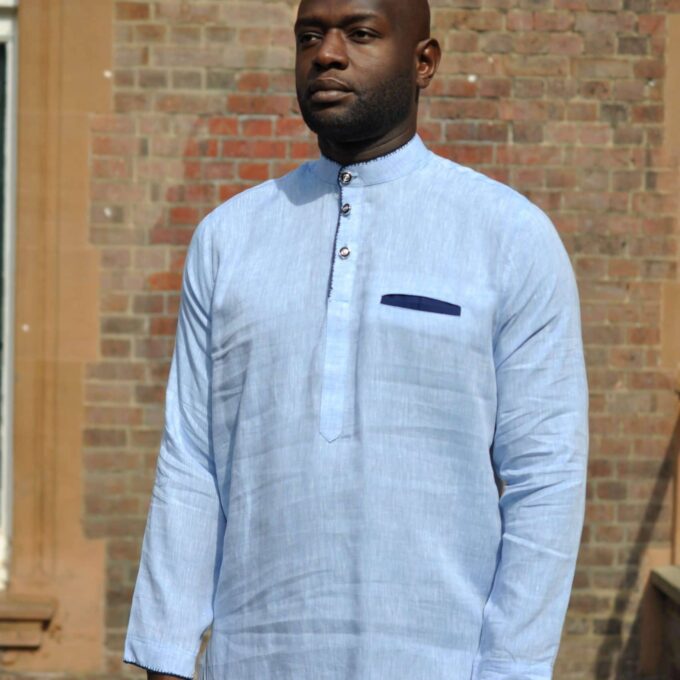 Frontal of model wearing a light blue men's long sleeve polo shirt made from linen. Features a front pocket with navy blue trim and overcast stitching on collar, cuffs and chest.