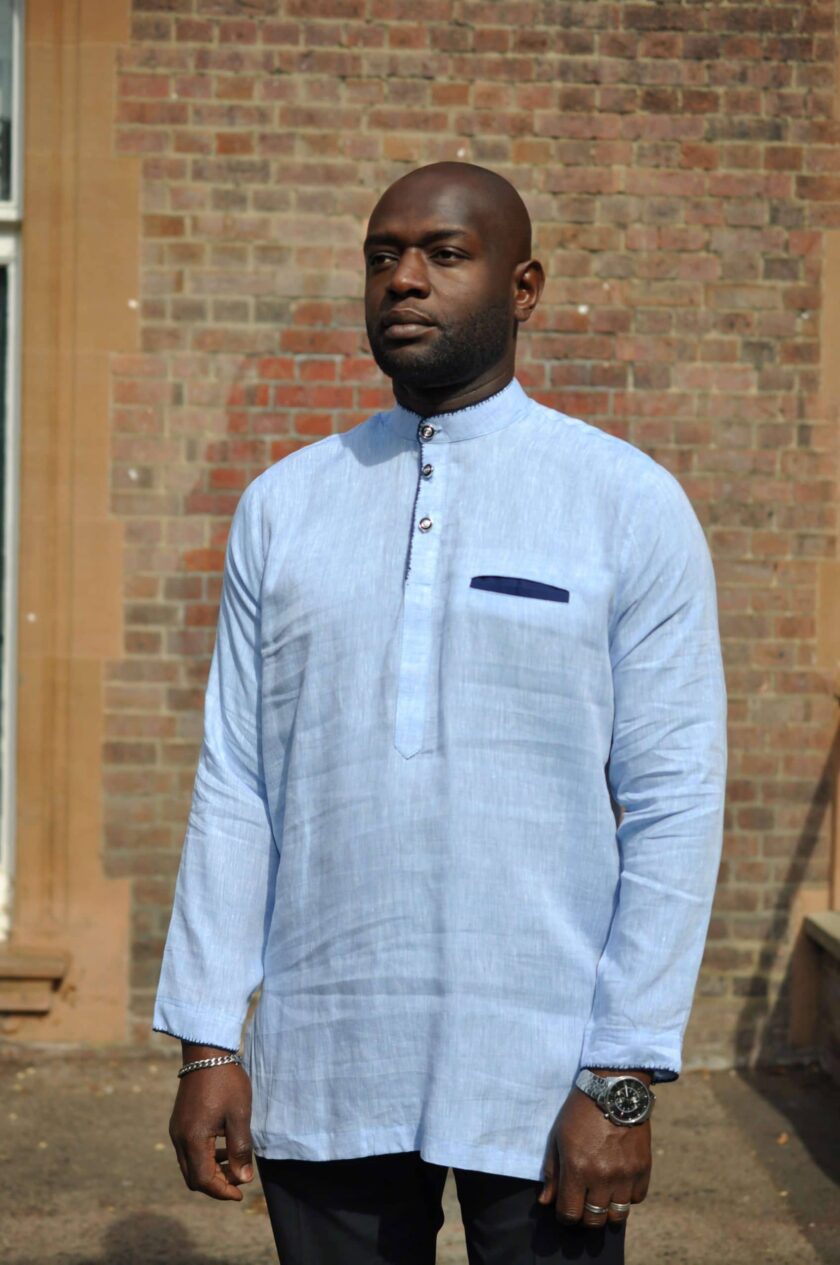 Frontal of model wearing a light blue men's long sleeve polo shirt made from linen. Features a front pocket with navy blue trim and overcast stitching on collar, cuffs and chest.