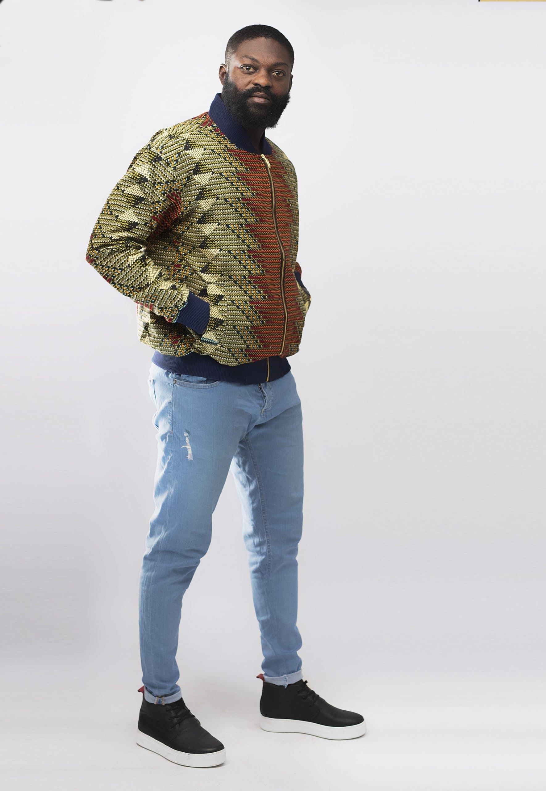 Side shot of model wearing our Abeba men's multi-coloured bomber jacket in all over African print pattern. Featuring 2 side pockets and a navy blue baseball collar and cuffs.