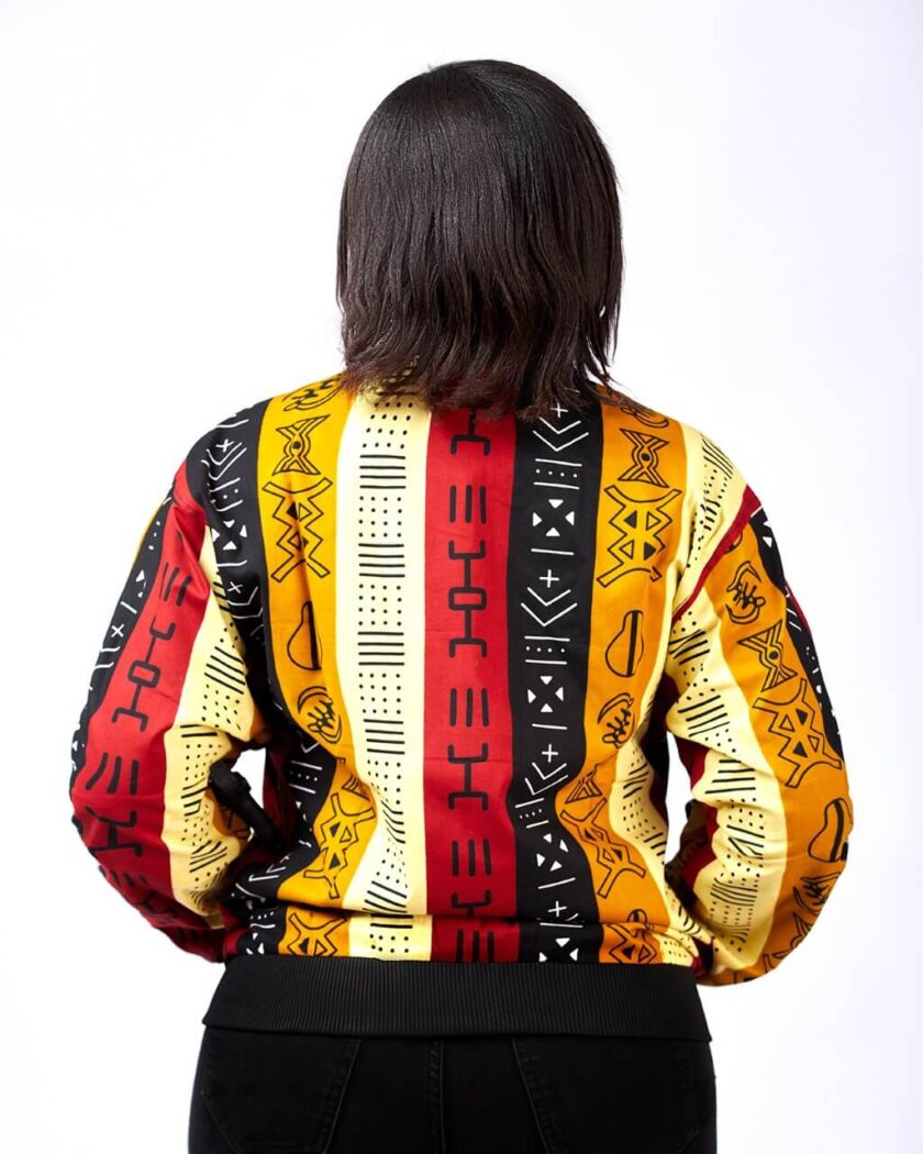 Back shot of model wearing a colourful, striped, ladies bomber jacket in all over African Kente print pattern.