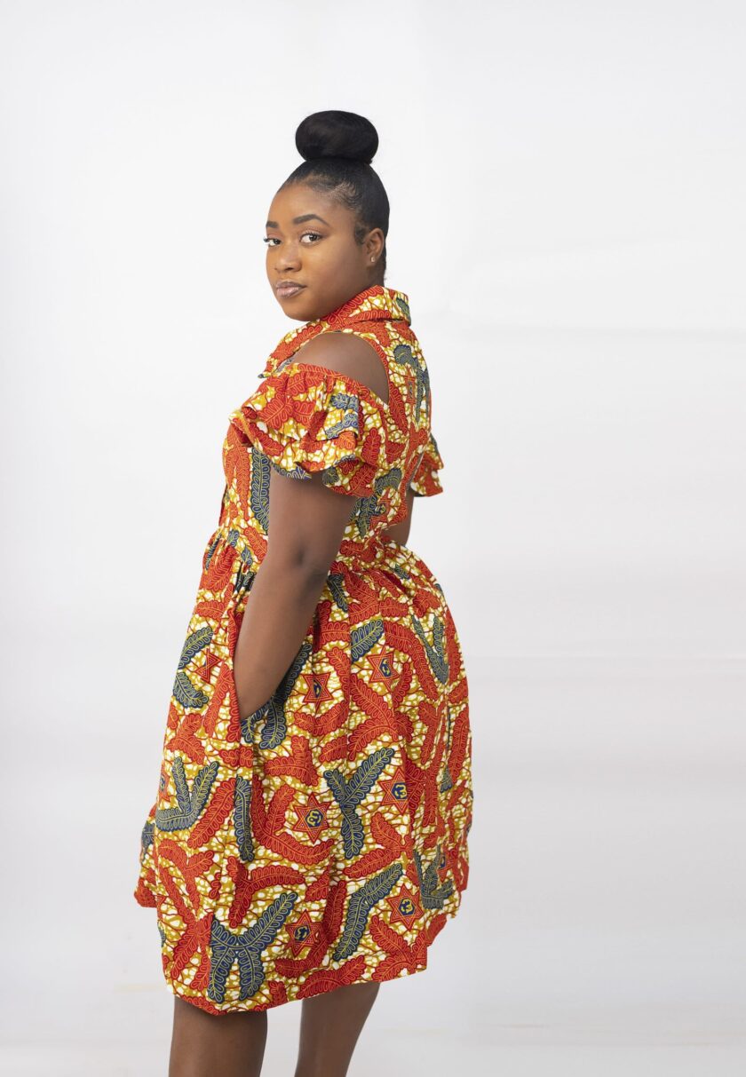 Side shot of model wearing our Oluchi skater midi dress in all over orange, gold and blue African print pattern. Features a cold shoulder, short sleeve, collar neckline.